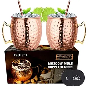 Two hammered copper mugs with straws and coasters on a rustic backdrop