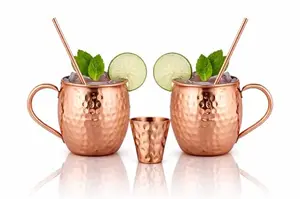 Two solid copper mugs with a shot glass and straws on a wooden board